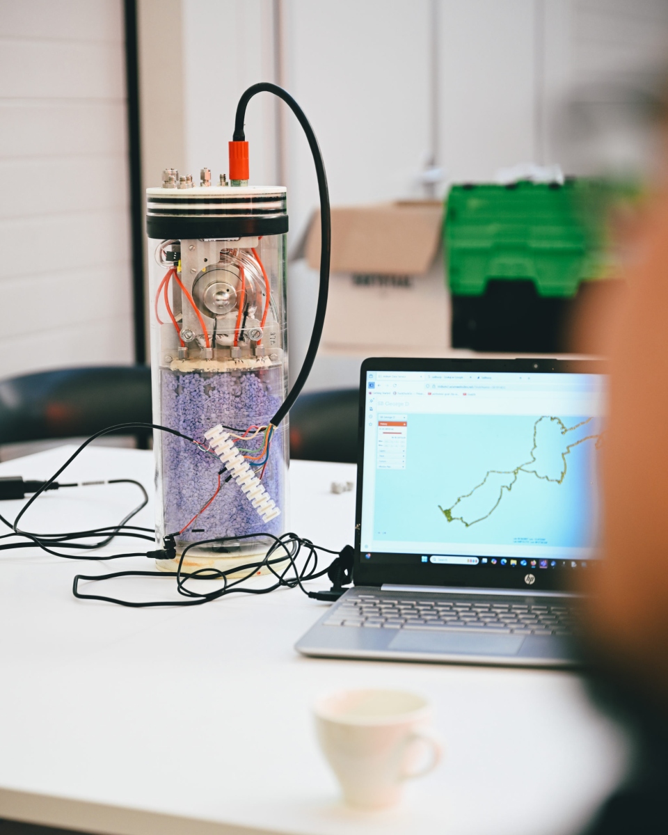 A cylindrical scientific instrument with a transparent casing, filled with purple desiccant beads and fitted with wiring and connectors, sits on a desk next to an HP laptop displaying a GPS track on a map.