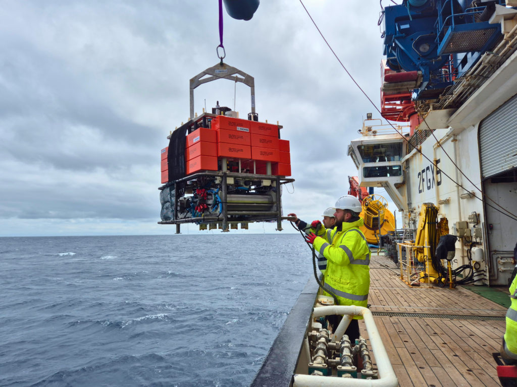 Crew members in high-visibility jackets and hard hats on the deck of a research vessel guide a large orange scientific instrument module — labelled GEORGE — as it is lifted by crane over the side of the ship