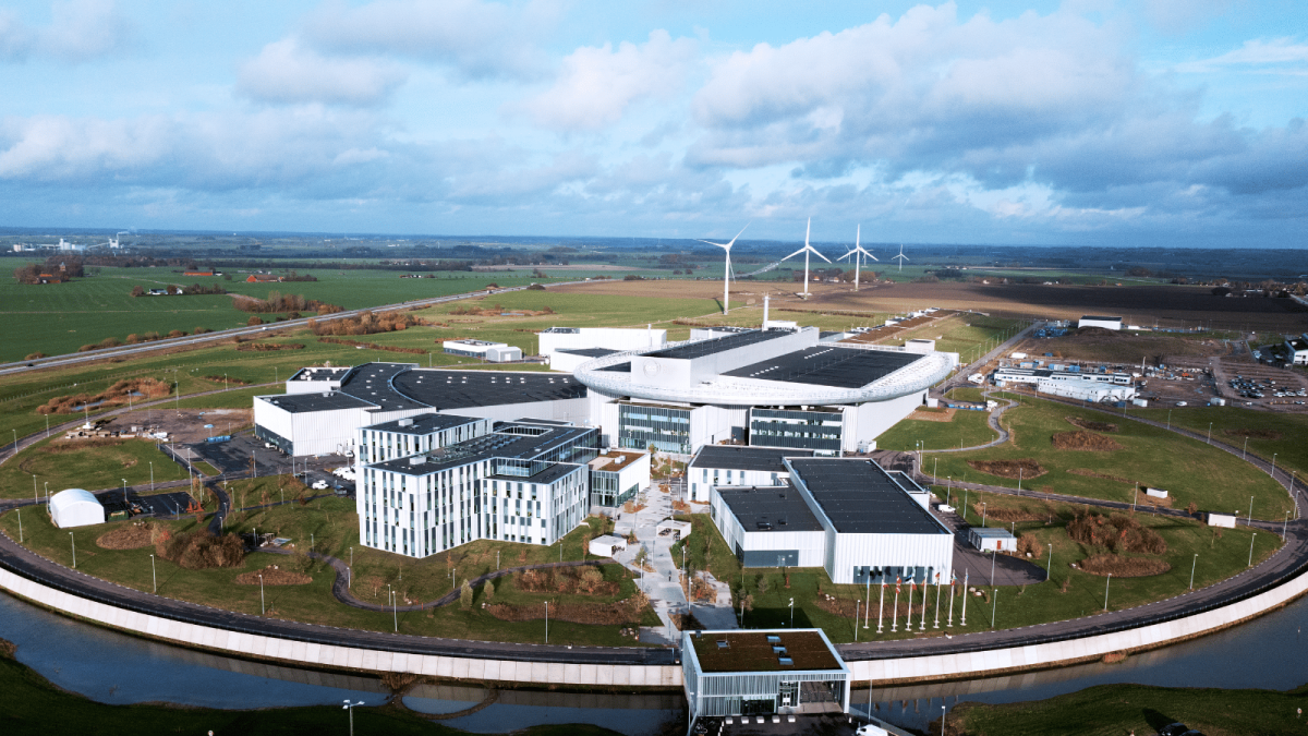 A large modern research facility with white and gray interconnected buildings arranged in a circular layout, surrounded by green fields and farmland. Wind turbines stand in the background, and a curved road borders the site.
