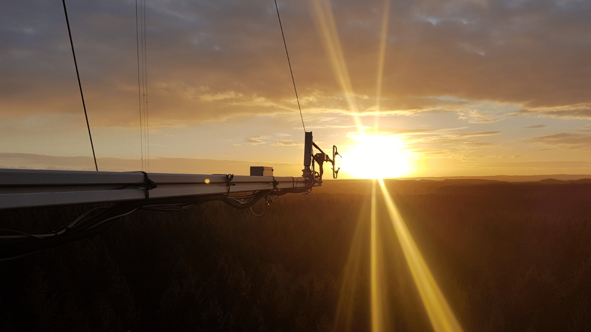 A dramatic sunset view from the top of a tall monitoring tower, with scientific instruments and sensor equipment silhouetted against a brilliant golden sun sitting low on the horizon. Rays of sunlight radiate outward across a vast, darkened landscape of forest stretching into the distance, beneath a warm orange and grey sky with wispy clouds.
