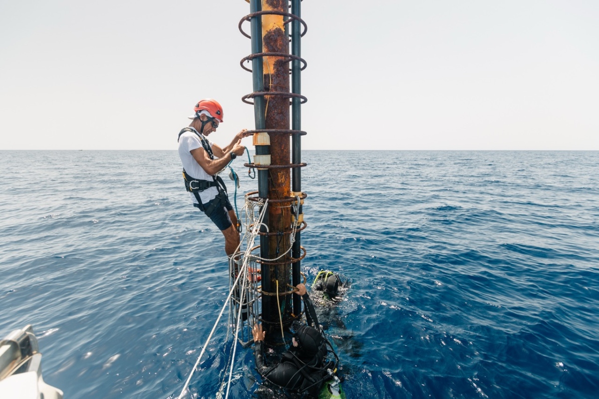 A helmeted technician in a safety harness works on an offshore monitoring mast while divers assist from the water below