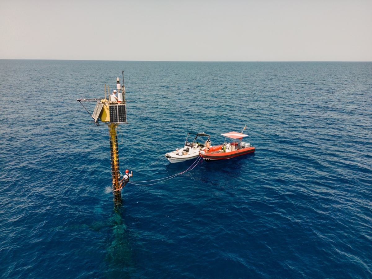 An offshore ocean-atmosphere monitoring station rises from the sea beside two small support boats, with a technician working on the structure.