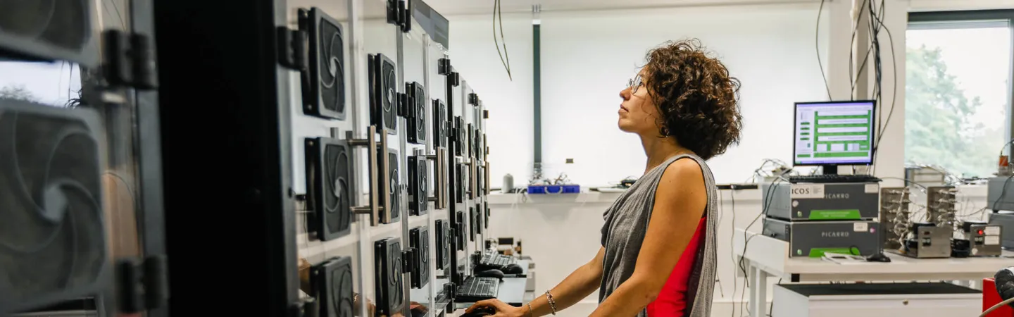 A person stands in a laboratory operating multiple computer monitors and scientific instruments mounted on a wall. The workspace includes labeled equipment, cooling fans, and neatly organized cables, creating a clean, high‑tech research environment.
