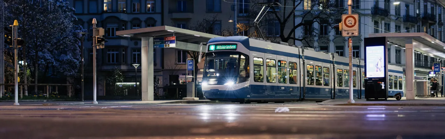 Electric tram at a city stop at dusk, with illuminated destination display and urban buildings in the background