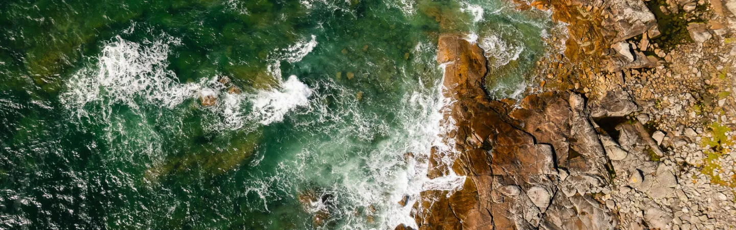 An aerial view of waves crashing against a rugged rocky coastline, with deep green ocean water on the left and jagged brown and gray rocks on the right.