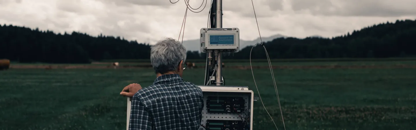 Researcher inspecting monitoring equipment in a grassy field under cloudy skies