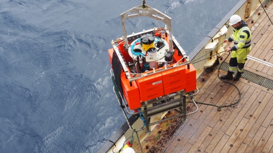 A large orange scientific instrument hangs from a crane beside a research vessel as crew members prepare for ocean deployment under cloudy skies