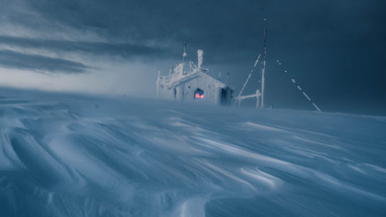 A small scientific monitoring station heavily encrusted with ice and snow stands in a blizzard on a barren, windswept arctic landscape. Drifting snow swirls across the foreground beneath a dark, stormy sky, while a warm red glow from the station's window and instruments provides a stark contrast to the harsh, frozen surroundings.