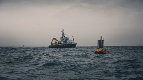 A research vessel equipped with yellow deck cranes sits on a choppy grey sea under an overcast sky. In the foreground, a yellow instrumented ocean monitoring buoy marked fitted with solar panels floats on the water’s surface.