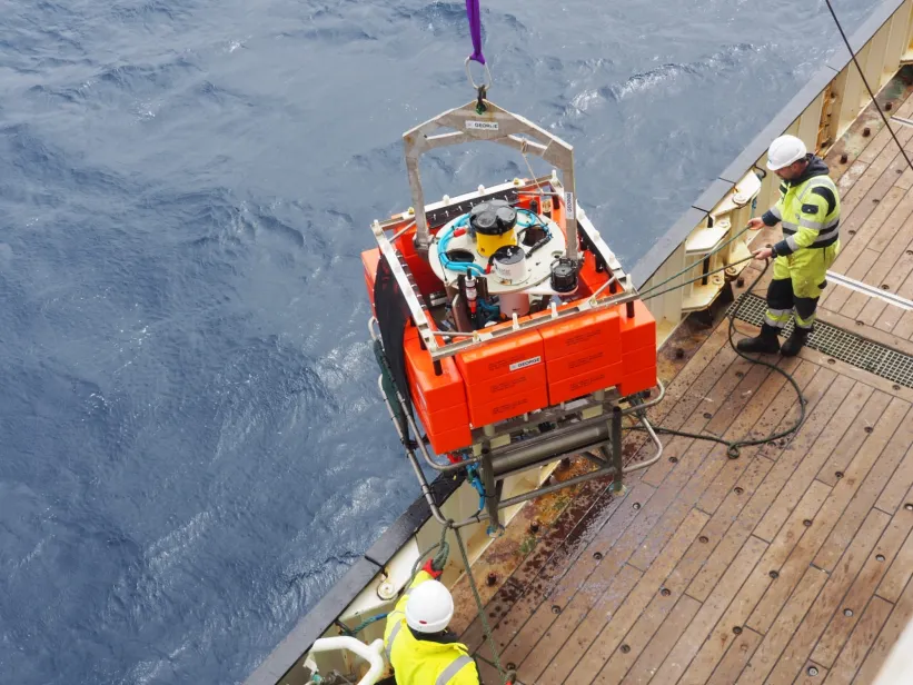 A large orange scientific instrument hangs from a crane beside a research vessel as crew members prepare for ocean deployment under cloudy skies
