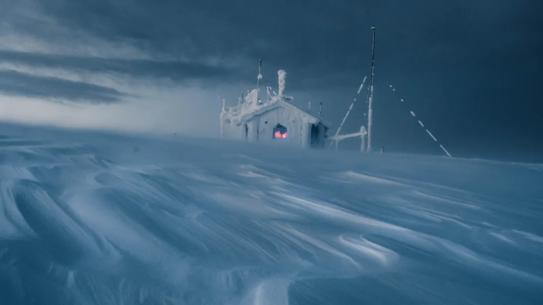 A small scientific monitoring station heavily encrusted with ice and snow stands in a blizzard on a barren, windswept arctic landscape. Drifting snow swirls across the foreground beneath a dark, stormy sky, while a warm red glow from the station's window and instruments provides a stark contrast to the harsh, frozen surroundings.