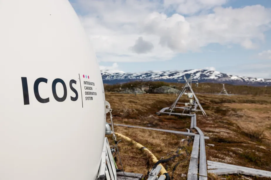 ICOS equipment at a tundra field site, with monitoring instruments extending across the landscape toward snow-covered mountains under a partly cloudy sky.