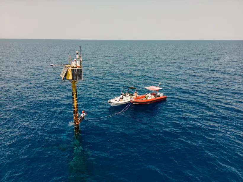 An offshore ocean-atmosphere monitoring station rises from the sea beside two small support boats, with a technician working on the structure.