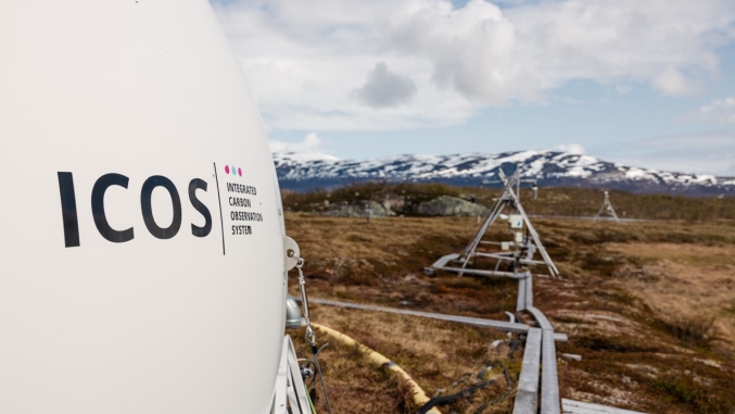 ICOS equipment at a tundra field site, with monitoring instruments extending across the landscape toward snow-covered mountains under a partly cloudy sky.