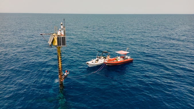 An offshore ocean-atmosphere monitoring station rises from the sea beside two small support boats, with a technician working on the structure.